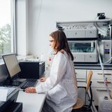 Eine Frau im weißen Kittel sitzt in einem Labor und schaut auf einen Bildschirm.