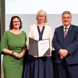 Drei Personen stehen festlich gekleidet nebeneinander, die Frau in der Mitte hält eine Urkunde in der Hand.