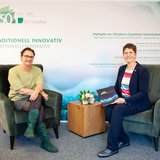 Zwei Frauen sitzen jeweils auf einem grünen Sessel, hinter ihnen ein bedrucktes Werbebanner.