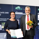 Eine Frau mit einer Urkunde in der Hand und ein Mann mit Blumen stehen vor einer Ausstellungswand.